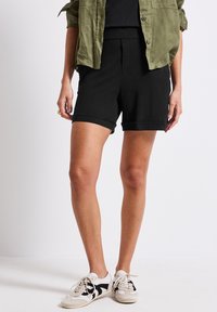 Shorts neri in cotone con orli arrotolati, abbinati a una maglietta scura e a una camicia button-up verde oliva. Sneakers bianche e nere con motivo.