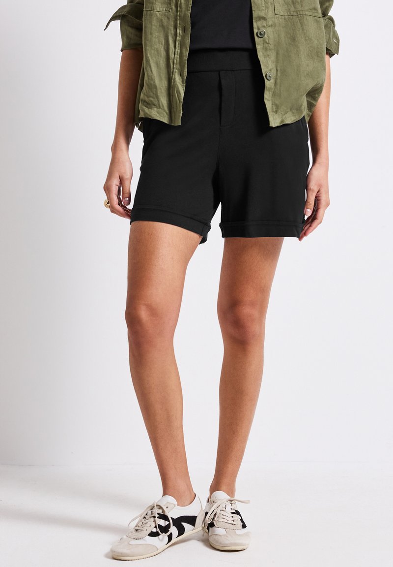 Shorts neri in cotone con orli arrotolati, abbinati a una maglietta scura e a una camicia button-up verde oliva. Sneakers bianche e nere con motivo.
