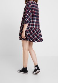 Femme portant une robe à carreaux rouge, blanche et bleu marine avec des manches retroussées et des baskets hautes Converse noires, debout sur un sol blanc.