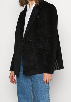 Persoon draagt een zwarte corduroy Adidas blazer over een wit overhemd en blauwe spijkerbroek, staand tegen een effen lichte achtergrond.