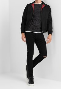 Veste zippée noire avec doublure en plaids rouges par-dessus un pull gris foncé, assortie à un jean noir déchiré et des baskets noires.