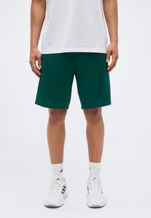 Mann trägt grüne Sportshorts, weißes T-Shirt, weiße Adidas-Socken und weiße Adidas-Sneakers, steht mit entspannten Armen neben dem Körper.
