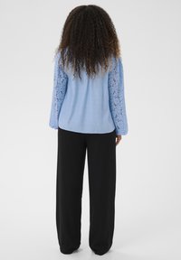 Blouse bleu clair avec manches en dentelle, associée à un pantalon large noir. La blouse est réalisée dans un tissu lisse avec des détails délicats en dentelle.