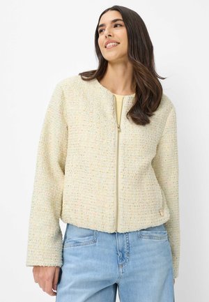 Vrouw die een crèmekleurig, gestructureerd jack met rits en lichtblauwe spijkerbroek draagt, glimlacht en iets zijwaarts kijkt tegen een witte achtergrond.