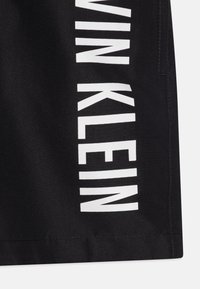 Pantalones cortos de tela negra con un texto de logo vertical en blanco "CALVIN KLEIN" en el lateral. Textura suave con corte recto y dobladillo cosido.