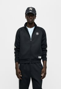 Μαύρο αθλητικό μπουφάν με φερμουάρ, ψηλό γιακά και λογότυπο στο στήθος· γκρι ρίγες στα πλάγια· συνδυασμένο με σκούρο καπέλο με κάθετες λευκές ρίγες.