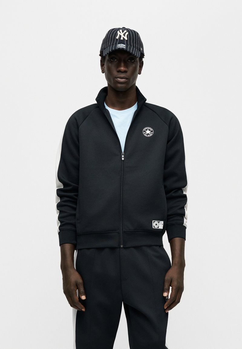 Sort trackjakke med lynlås, høj krave og logo på brystet; grå sidestriber; kombineret med en mørk kasket med lodrette hvide striber.