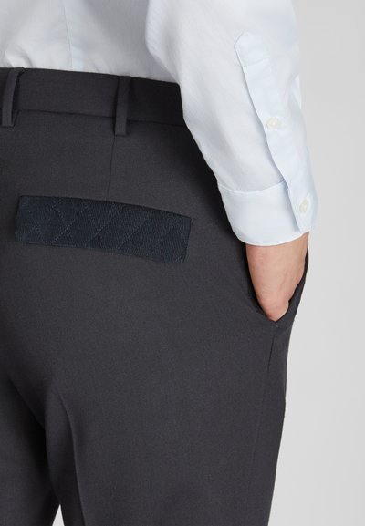 Donkergrijze op maat gemaakte broek met een gestructureerd marineblauw label op de achterzak, gemaakt van een gladde stof. Mouwen van een overhemd zichtbaar boven de tailleband.