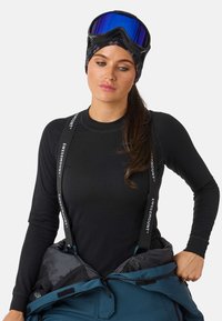 Schwarzes, geripptes Langarmoberteil mit verstellbaren Trägern und türkisfarbenen Ski-Hosen. Blaue, verspiegelt Sonnenbrille und ein gemusterter, schwarzer Haarreif runden den Look ab.