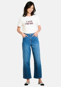 Biela tričko s potlačou "CAFE CREME" v burgundskej farbe, skombinované s modrými vysoko pásovými širokými džínsami. Model stojí so zapichnutými rukami v vreckách.