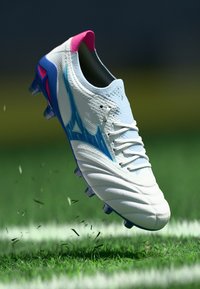 Călcâi de fotbal alb cu accente roz și bleu, suprafață texturată, design cu șireturi și mai multe tinte pe talpă, plutind deasupra ierbii.
