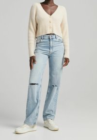 Krémový pletený krátký kardigan na knoflíky, v kombinaci se světle modrými džíny s vysokým pasem s roztrhanými detaily a bílými teniskami.