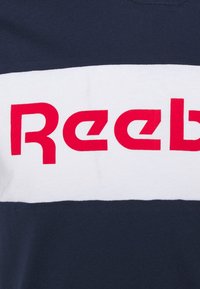 Marinblå bomullströja med ett horisontellt vitt rand och fet röd text "Reebok". Mjuk textur, korta ärmar och rundad halsringning.