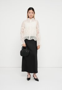 Weiße Spitzentbluse mit langen Ärmeln und hohem Kragen, kombiniert mit einem schwarzen Maxirock. Das Modell trägt eine kleine strukturierten schwarze Handtasche.