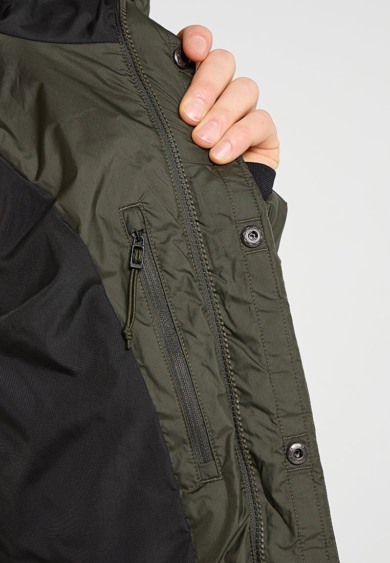 OlivgrüneJacke mit schwarzen Akzenten; verfügt über einen Reißverschluss vorne, Druckknöpfe und eine Textur, die weich und isoliert wirkt.