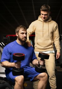 Deux personnes dans une salle de sport. L'une tient des haltères noirs avec des accents rouges, portant une tenue de sport bleue ; l'autre porte un sweat à capuche beige et un pantalon.