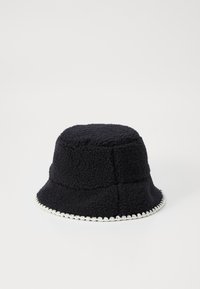 Faux fur svart bøttehatt med en teksturert overflate, i tillegg til en kontrasterende hvit kant langs kanten. Sylinderformet, mykt materiale.