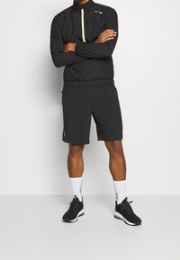 Černá sportovní bunda s zelenými zipovými detaily, spojená s černými šortkami. Model nosí černé sportovní boty a bílé ponožky.