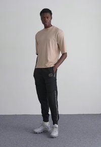 Beżowy t-shirt z krótkim rękawem w połączeniu z czarnymi spodniami cargo z bocznymi paskami i kieszeniami, uzupełniony szarymi butami sportowymi.
