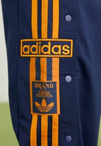 Mørkeblå bukser med orange Adidas-logo, tre lodrette striber og trykknapper langs sidesømmen.