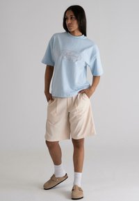 Light blue cotton t-shirt with logo, paired with beige pleated shorts. Model wears white socks and tan slip-on shoes. Neutral background.