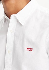 Camicia bianca con bottoni realizzata in cotone, dotata di un colletto a punta e caratterizzata da un piccolo logo rosso Levi's sul petto. Texture morbida e design classico.