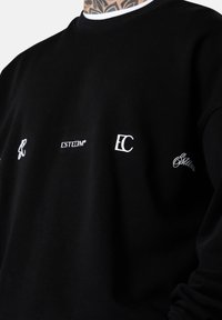 Sweatshirt noir en tissu doux, arborant des logos blancs : "K," "ESTEEM," "EC," et un accent en script. Col et poignets côtelés.
