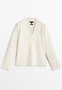 Pull en maille côtelée blanche avec un décolleté en V, des manches longues et une coupe ample. Le tissu présente une finition texturée, avec une silhouette légèrement décontractée.
