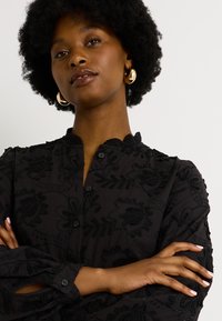 Blusa negra de botones con bordado floral texturizado, cuello mandarín y mangas largas con detalles fruncidos.