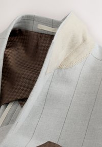 Límec šedého látkového saka s béžovým akcentem; vnitřní část má vzor hnědého pepita. Hladká textura s viditelnými detaily stehu.