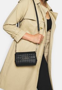 Nainen, jolla on beige trenchcoat ja musta mekko, pitää mustaa vuorattua Guess-käsilaukkua, jossa on ketjuolkaimet olkapäällä.