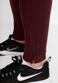 Close-up van bordeauxrode sportbroeken met enkelrits, gedragen met zwarte Nike sneakers en witte sokken op een witte achtergrond.