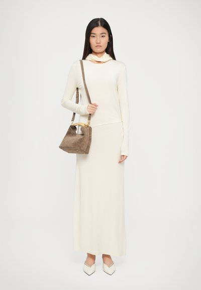 Giovane donna che indossa un lungo vestito crema e scarpe abbinate, mentre tiene una borsa in camoscio taupe con manico superiore e tracolla.