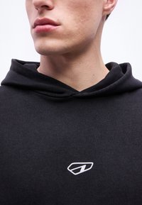 Κοντινό πλάνο του κάτω μέρους του προσώπου και του λαιμού ενός ατόμου που φοράει μαύρη μπλούζα με κουκούλα, με ένα μικρό λευκό γεωμετρικό λογότυπο στο στήθος.