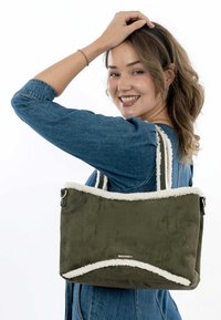 Borsa a spalla verde oliva con bordi bianchi pelosi, dotata di doppi manici e una superficie morbida simile alla pelle scamosciata. Logo visibile sul davanti.