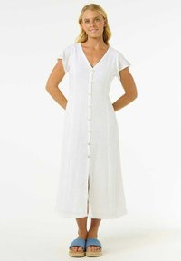 Robe maxi blanche à boutons avec des manches courtes en volants, en tissu texturé. Portée avec des sandales bleues, présentant un design simple et élégant.