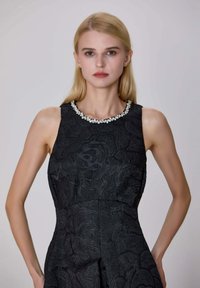 Jeune femme avec des cheveux blonds raides portant une robe sans manches noire texturée avec un col orné de bijoux devant un fond uni.