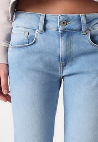 Šviesiai mėlynos džinsinės kelnės su aukštu liemeniu, penkių kišenių dizainu ir kontrastingais geltonais siūlais. Turi užtrauktuką ir sagą.