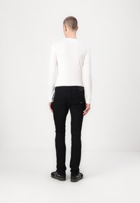 Una persona con el cabello corto está de pie frente a una pared blanca, vistiendo una camiseta blanca de manga larga, pantalones negros y zapatillas negras con detalles blancos.