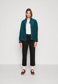 Γυναίκα φοράει μπλε κοτλέ πουκάμισο πάνω από λευκό τοπ, μαύρο κοντό παντελόνι, λευκές κάλτσες και μπορντό αθλητικά παπούτσια σε απλή φόντο.