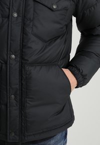 Sort puffjakke med trykknapper, to frontlommer, elastiske manchetter og en glat, quiltet struktur, der har et klassisk design og silhuet.