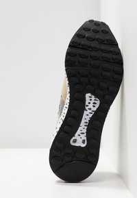 Semelle de basket avec des crampons circulaires noirs et une semelle intermédiaire blanche parsemée de taches noires irrégulières, inclinée contre un mur et un sol blancs.