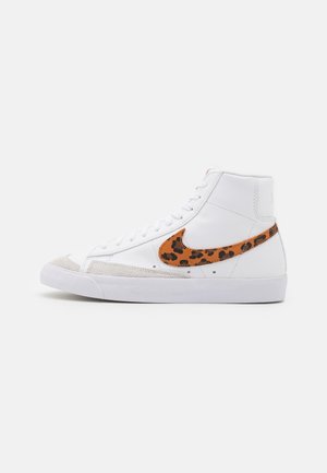 Biały sneaker z wysoką cholewką, zamszowym noskiem i brązowym logo Nike z czarnym wzorem w cętki.