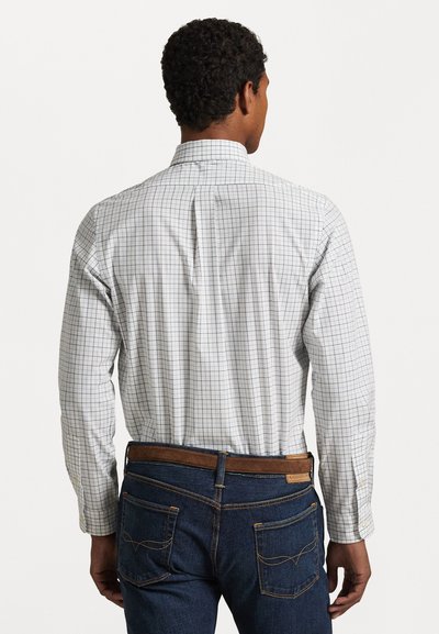 Camisa blanca de manga larga con un patrón de lunares azul y gris, con cuello abotonado y pliegue en la parte posterior, combinada con jeans de mezclilla oscura.