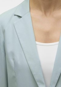 Blazer azzurro chiaro con rever strutturato, texture del tessuto liscia e vestibilità slim, abbinato a un top bianco sottostante.
