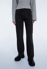 Pantalon cargo noir en coton, avec une coupe ample, deux poches latérales et des coutures épaisses. Associé à un pull gris texturé et des bottes noires épaisses.