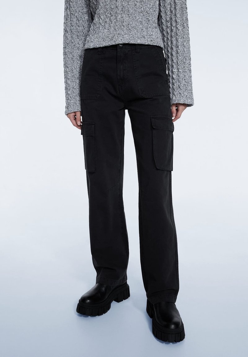 Pantalon cargo noir en coton, avec une coupe ample, deux poches latérales et des coutures épaisses. Associé à un pull gris texturé et des bottes noires épaisses.