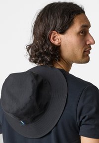 Black bucket hat made from lightweight fabric, featuring a wide circular brim and a blue and yellow accent detail. Worn at an angle on the back.