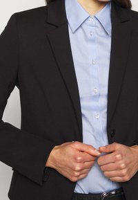 Blazer nero su misura sopra una camicia a collare azzurro chiaro, con chiusure a bottone, una consistenza liscia e maniche strutturate.