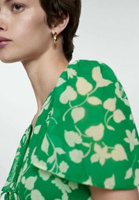 Rochie cu model floral verde, din material transparent, cu mâneci puff scurte și detaliu legat la decolteu. Cercei rotunzi aurii sunt purtați.